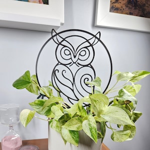 May include: A white ceramic plant pot with a green and white plant, and a black metal owl decoration. The owl is within a circular frame. A candle and hourglass are on a wooden surface.