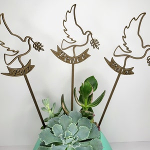 May include: Three wooden dove figurines with olive branches in their beaks, each with a banner reading "Peace", "Paz", and "Pax". The doves are arranged in a succulent planter.