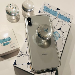 May include: A clear phone case with a pop socket featuring a blue butterfly design. The phone case is labeled "iPhone". The pop socket is on a white and blue background with the text "Triki Shop" in a bubble font.