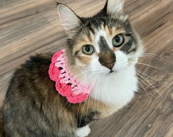 May include: A calico cat with green eyes wearing a handmade crochet collar. The collar features a scalloped edge design with alternating layers of light pink and vibrant hot pink yarn, creating a textured, ruffled appearance around the neck.
