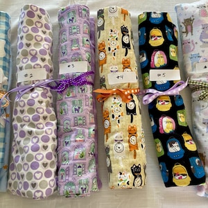 May include: Six rolled fabric pieces labeled #1 through #6. #1 has blue gingham and cats, #2 has purple circles, #3 has cats in windows, #4 has cats and clocks, #5 has cats in backpacks on black, and #6 has colorful cats on white fabric.