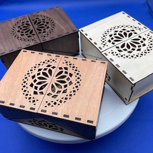 Puede incluir: Tres cajas de madera con diseños florales cortados con láser en las tapas. Las cajas son en tonos marrón y madera natural, con una base oscura. Las cajas se exhiben sobre una superficie blanca sobre un fondo azul.