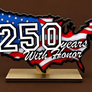 May include: A decorative sign shaped like the map of the United States featuring the American flag pattern. The sign displays the large white text 250 and the script text years With Honor. It is mounted on a rectangular metallic base stand.