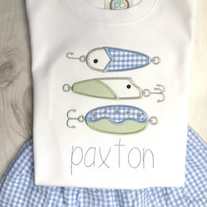 May include: White long-sleeved shirt with three embroidered fishing lures in blue and green gingham patterns. The name "paxton" is embroidered in blue script below the lures. The shirt is paired with a blue and white gingham skirt.