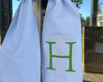 May include: A light blue scarf with an embroidered green and white monogram letter "H". The scarf is tied to a green wreath. The scarf is made of a soft fabric and hangs vertically.