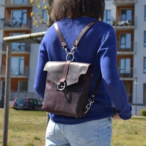 Puede incluir: Una mochila de cuero marrón con herrajes plateados y correas ajustables. La mochila la lleva puesta una persona que lleva un jersey azul y unos vaqueros azules.