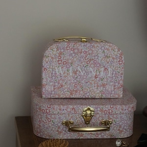 May include: Two floral patterned decorative boxes, one stacked on top of the other. The top box has the name "WINNIE" printed on it. Both boxes have gold-coloured handles and clasps. A wooden hairbrush is on the table.