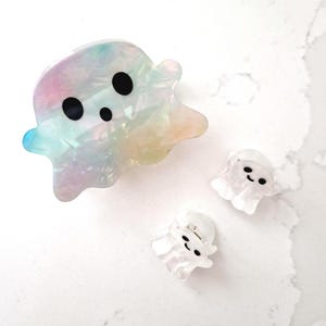 Op de afbeelding: Een grote iriserende spookvormige haarclip met drie zwarte stipogen en twee kleinere doorzichtige spookvormige haarclips met zwarte stipogen. De clips liggen op een wit marmeren oppervlak.