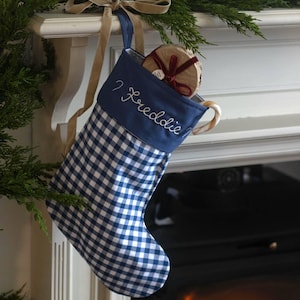 May include: A blue and white gingham Christmas stocking with a solid blue cuff featuring the embroidered name Freddie in white script. The stocking includes a beige ribbon loop for hanging and contains a small round gift wrapped in brown paper.