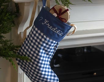 Op de afbeelding: Een blauw-witte gingham kerstkous met een effen blauwe boord waarop de naam Freddie in witte schrijfletters is geborduurd. De kous heeft een beige lintlus om op te hangen en bevat een klein rond cadeautje verpakt in bruin papier.