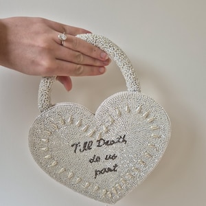 Puede incluir: Un bolso en forma de corazón, con cuentas blancas, con un asa con cuentas. El bolso presenta el texto "Till Death do us part" en escritura negra. El bolso es sostenido por una mano con un anillo de diamantes.