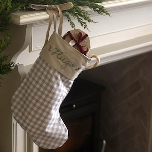 Op de afbeelding: Een beige en witte kerstkous met vichy-ruitjes en een crèmekleurige boord waarop de naam Hugo in groene schrijfletters is geborduurd. De kous hangt aan een beige lint en bevat een rond cadeau verpakt met een rood fluwelen lint.