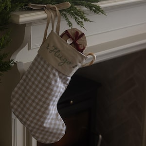 Puede incluir: Un calcetín navideño a cuadros beige y blanco con el nombre "Hugo" bordado en verde. Cuelga de una repisa blanca. Dentro hay un regalo envuelto con una cinta roja. La repisa está adornada con vegetación y un lazo.