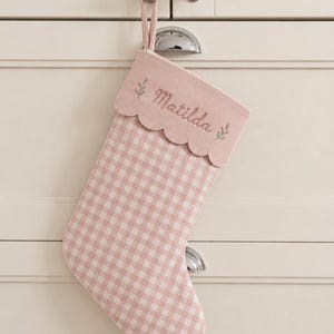 May include: A pink and white gingham Christmas stocking featuring a solid pink scalloped cuff. The cuff is embroidered with the name Matilda in a cursive script, flanked by small green leaf sprigs on either side. A matching pink fabric loop is attached.
