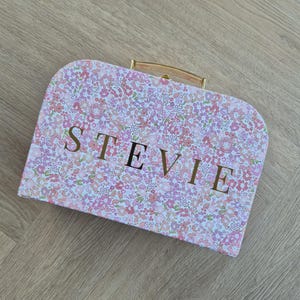 May include: A pink floral patterned suitcase with the name "STEVIE" in gold lettering. The suitcase has a gold handle and clasp. The suitcase is on a light brown wooden surface.