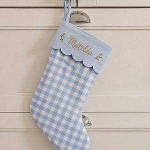 Könnte beinhalten: Ein hellblau-weiß karierter Weihnachtsstrumpf mit einer gewellten Manschette, die mit dem Namen Matilda in Goldfaden bestickt ist, akzentuiert durch kleine Blumenmotive auf beiden Seiten und einer passenden hellblauen Schlaufe.