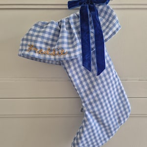 May include: A light blue and white gingham Christmas stocking featuring a folded cuff embroidered with the name Freddie in gold thread. The stocking is accented with a deep blue velvet ribbon tied into a bow at the top hanging loop.