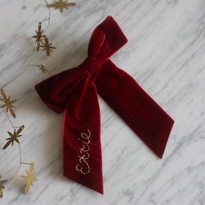 May include: A rich burgundy velvet bow, embroidered with the name "Exxie" in gold thread. The bow is neatly tied with long tails. A gold star garland is visible in the background. This could be a hair accessory or a decorative item.