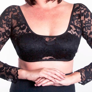 Könnte beinhalten: Schwarzes Spitzen-Crop-Top mit langen Ärmeln. Das Oberteil hat einen runden Ausschnitt und zarte florale Spitzendetails. Die Ärmel haben einen leichten Schlag am Handgelenk. Das Kleidungsstück kann als Layering-Piece oder als Einzelteil getragen werden.