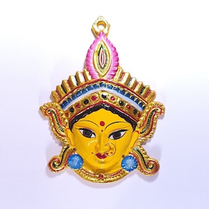 May include: A gold-coloured metal pendant featuring a colourful face with a crown. The face is yellow with black hair, red and black markings, and blue eyes. The crown is decorated with gold, blue, red, and white details.