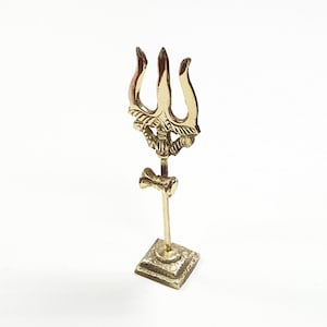 Puede incluir: Una estatua de tridente de metal dorado sobre una base cuadrada. El tridente tiene tres puntas y detalles intrincados.