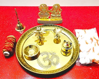 May include: A gold metal plate with a raised Om symbol in the center. The plate is surrounded by a decorative border and contains a variety of small gold metal items, including a diya, a small incense burner, and a flower-shaped object. There are also cotton wicks and a spool of red and gold thread.