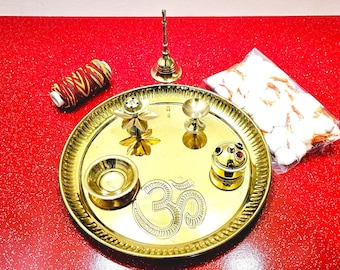 May include: A gold metal plate with a raised Om symbol in the centre. The plate is surrounded by a variety of small gold metal items, including a bell, a diya, a flower-shaped incense burner, and a small bowl. The items are arranged on a red background.