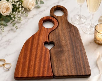 May include: Two wooden serving boards with circular handle cutouts, one in a lighter reddish-brown wood and one in a darker brown wood, positioned together so that a heart-shaped cutout is formed where the two boards meet at their curved edges.