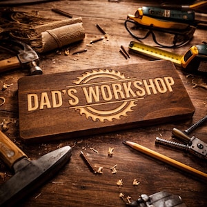 May include: A wooden sign with the text "DAD'S WORKSHOP" is surrounded by tools like hammers, a measuring tape, and safety glasses. The sign has a circular saw blade design around the text, and the tools are arranged on a wooden surface.