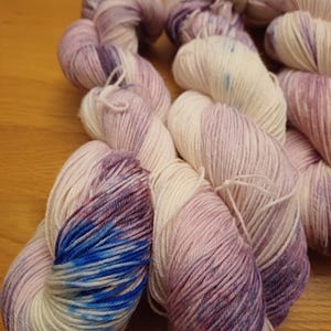 May include: Three skeins of yarn in shades of white, pink, and purple with blue accents. The yarn is hand-dyed and has a variegated colour pattern.