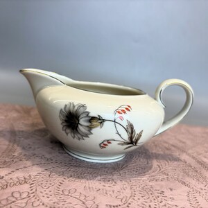 May include: A white porcelain creamer with a floral design. The creamer has a gold rim and a delicate floral pattern in shades of gray, red, and green.