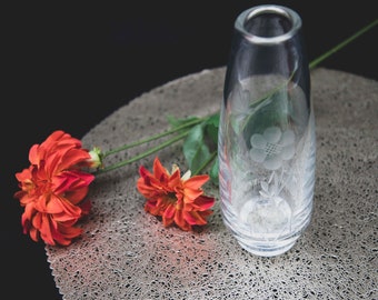 Op de afbeelding: Een heldere glazen vaas met een gegraveerd bloemenmotief staat op een zilverkleurig, getextureerd oppervlak. Twee oranje bloemen liggen op het oppervlak naast de vaas.