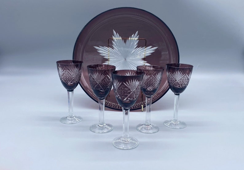 Pu&ograve; includere: Set di sei bicchieri da liquore a stelo in vetro viola con un design in vetro tagliato. I bicchieri sono disposti a semicerchio attorno a un vassoio da portata in vetro viola con un design in vetro tagliato.