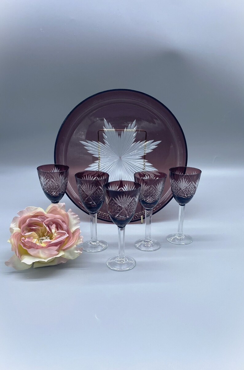 Pu&ograve; includere: Set di sei bicchieri da vino in vetro viola con un design in vetro tagliato. I bicchieri sono disposti a semicerchio su una superficie bianca con un vassoio da portata in vetro viola dietro di loro.