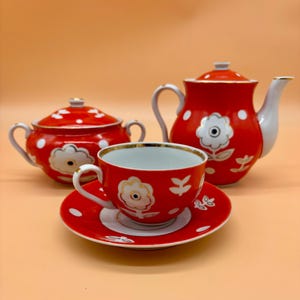 May include: A red and white floral tea set, including a teapot, sugar bowl, teacup, and saucer. The set features a red background with white polka dots and floral designs. The teacup sits on a matching saucer.