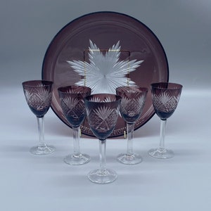 Pu&ograve; includere: Set di cinque bicchieri da vino in vetro trasparente con una tinta viola e disegni intricati tagliati. I bicchieri sono disposti a semicerchio attorno a un grande piatto da portata in vetro tagliato viola.