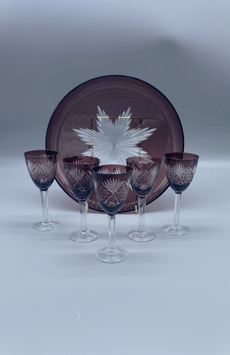 Pu&ograve; includere: Set di cinque bicchieri da vino in vetro trasparente con una tinta viola e disegni intricati tagliati. I bicchieri sono disposti a semicerchio attorno a un grande piatto da portata in vetro tagliato viola.