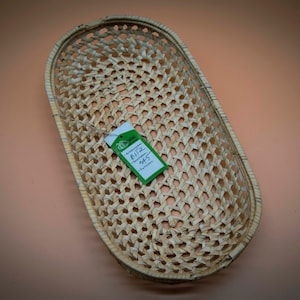 Può includere: Un cesto ovale intrecciato in materiale naturale. Il cesto è di colore marrone chiaro e presenta un motivo intrecciato. Un'etichetta verde con il testo "Basket 81/2" 945" è attaccata al cesto.