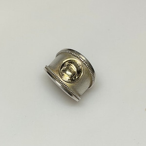 Puede incluir: Un anillo de plata con un diseño dorado en el centro. El anillo tiene una banda texturizada.