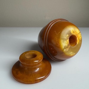 May include: A vintage, amber-coloured, turned wood container with a matching lid. The container has a rounded, bulbous shape with a central hole and a lid with a small, circular opening. The wood grain is visible, and the finish is glossy.