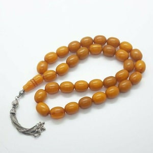Puede incluir: Un collar de cuentas de oración de ámbar con un borla de plata. Las cuentas son de un color ámbar rico y cálido y tienen forma ovalada. La borla es de plata y tiene un diseño decorativo.