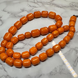 May include: A long strand of orange amber beads, strung together to form a necklace. The beads are all roughly the same size and shape, and they have a smooth, polished finish.