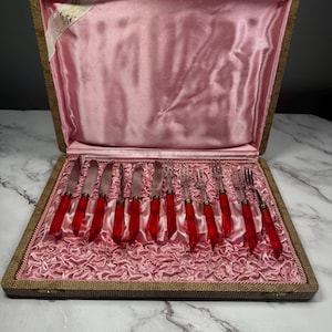 Peut inclure: Un ensemble de couverts vintage avec des manches en plastique rouge dans un étui en similicuir marron avec une doublure en satin rose.