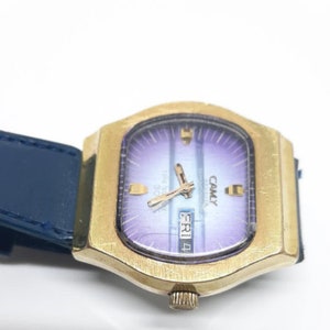 May include: A vintage gold-toned CAMY watch with a rectangular face and a blue leather strap. The watch face has a purple and blue gradient with gold hands and markers. The day and date are displayed. The watch is set to Friday, the 4th.