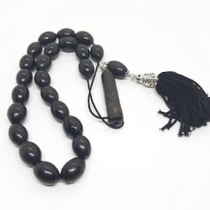 Peut inclure: Un mala de prière noir avec un pompon et un charme argenté. Les perles sont de forme ovale et le pompon est noir.