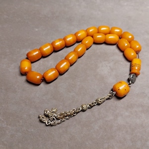 May include: A string of amber prayer beads with a silver tassel and a silver charm. The beads are a rich, warm amber colour and are strung on a thin cord. The tassel is made of silver beads and is attached to the end of the string. The charm is a small, silver disc with a design on it.