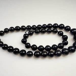 Może przedstawiać: Długi sznur polerowanych czarnych koralików ułożonych w podwójną pętlę. Koraliki są kuliste i różnią się wielkością, przy czym największy koralik znajduje się blisko końca sznura. Powierzchnia każdego koralika jest gładka i odblaskowa z białymi refleksami.