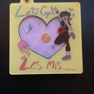 May include: A yellow and pink heart-shaped keychain featuring a cartoon character holding a guitar. The text "Let's Go!" and "Les Mis World Tour" are on the keychain.