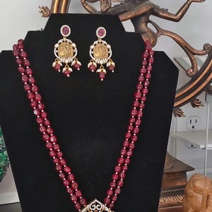 May include: A maroon beaded necklace and matching earrings set. The necklace features a large pendant with a gold-toned center and a detailed design. The earrings have a similar design. The jewelry is displayed on a black stand.