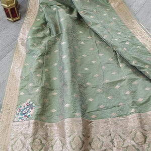 May include: A sage green saree featuring a wide metallic gold border with intricate floral patterns. The fabric has a subtle sheen and is decorated with small, repeating gold motifs throughout. A colorful floral patch is visible near the hem.